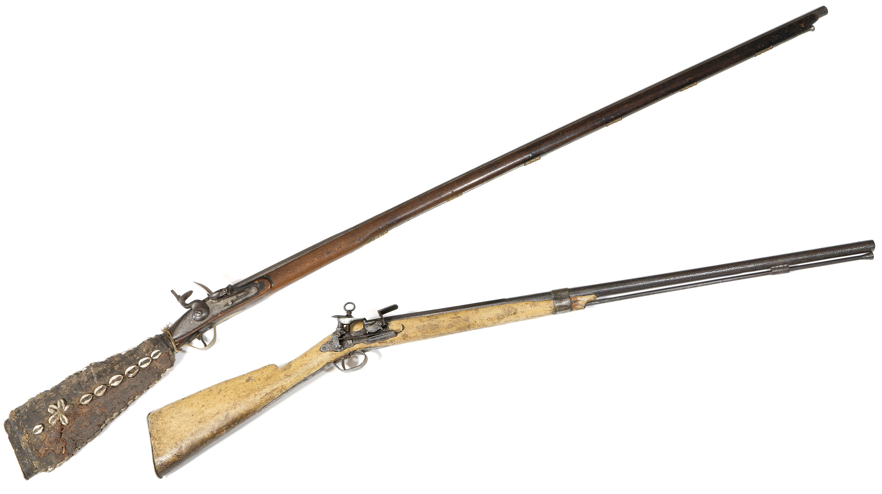 Trade guns. A large rifle, almost two metres long, with a dark wooden body and steel barrel and mechanism. The butt is coated in a fibrous material and is decorated with strings of cowrie shells. Also a flintlock rifle with a light wooden butt and handle, with a steel mechanism and barrel.