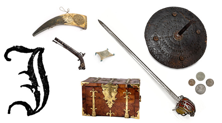 A large, ornate printed capital letter J with a box, a sword, pistol, shield, horn, cushion and medals.