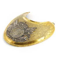 Gorget. A large, gold crescent shaped plate decorated with leaves around the edges. It has a silver stamp with knotwork and a crown surrounded by cannons, axes and swords.