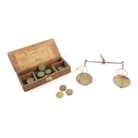 Guinea scales. A small set of scales consisting of two brass pans hung on either end of a rod with a hinge in the centre. There is also a box containing thick brass weights resembling coins.