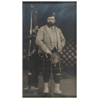 Photograph of Edward VII. A large bearded white man wearing a black cap, tartan jacket and waistcoat, a shirt and tie, a tartan kilt with a small swastika on it, a sporran, stockings and shiny black leather shoes, holding a walking stick.