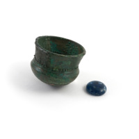 Grave goods. A miniature bronze cauldron covered with green rust and decorated with punched dots, and a small opaque blue glass object shaped like a lens.