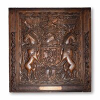 Heraldic panel. A large square dark wooden panel, about a metre wide, carved with a coat of arms containing a lion, held aloft by two chained unicorns with crowns around their necks. Above the coat of arms is a knight&#039;s helmet with a crown, and a lion holding a sword and flag on top. The background is filled with oak leaves.