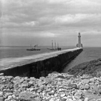 South Breakwater, Aberdeen around 1920. A concrete breakwater with a tower at the end projects from a rocky shore. Two figures stand on the breakwater in the distance. A small steamboat and a sailboat head past the breakwater, and the Aberdeenshire coast is visible on the horizon.