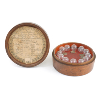 Glass bubbles. A set of twelve small glass bubbles each with a number engraved on them, in a circular wooden box lined with red fabric. The inside of the lid has a paper label which says GLASS BUBBLES, For proving spirits, Made by CAESAR ALTRIA, Mathematical Instrument Maker, ABERDEEN. A table with the numbers on the bubbles corresponding to strengths of alcohol is underneath.