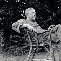 Photograph of Alexander Ogston relaxing in a wicker chair outdoors. He is seen looking into the distance and the photograph taken from side view. He reclines in a wicker chair with his leg crossed and his arm on th chair. 