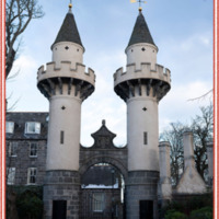 Powis Gateway. A gateway composed of a grey granite masonry arch between two medieval style towers with masonry on the lower portion and beige stucco on the rest. The towers have conical roofs with weather vanes in the shape of a crescent moon on its back.