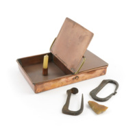 Tinder box. A copper box with two lid panels. In one half is a candle holder which has a candle in it. Next to the box are two buckle shaped pieces of metal and a piece of flint.