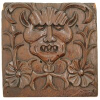 Panel. A wooden panel about twenty centimetres wide, carved with the face of a horned devil with pointy ears and teeth, with flowers and stems on either side.