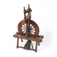 Spinning wheel. A wooden apparatus almost a metre tall, consisting of a frame with a pedal at the bottom, a large wheel with decoratively turned spokes, and a device at the top with a spindle and small metal hooks.