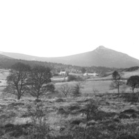 Bennachie. A mountain with a rounded peak, slightly snow covered. In the mid distance are two houses, fields and trees.