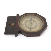 Table compass. A compass, eleven centimetres wide, in an octagonal wooden frame with a glass cover bordered by brass. The compass needle sits above a disc of paper printed with an anthropomorphic sun in the centre and directions and degrees around the edge.