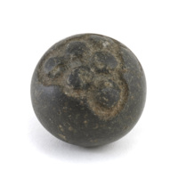 Carved stone ball. Roughly the size of a tennis ball. Black stone ball, very round and polished smooth except for a triangular shaped depression on one side which has six shallow round knobs arranged in a pyramid pattern.