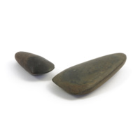 Stone axe heads. Two polished grey stone axe heads, one about four inches long, the other about seven inches long. The smaller one has a curved blade edge and is a round tapering oval shape, the larger has a straight edge and is a rounded triangular shape.