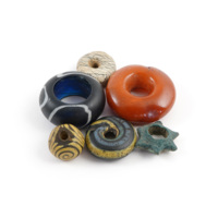 Beads. Six small beads, clockwise from top: a white ring with black veins made of steatite, an orange ring of amber, a blue star shaped bead, a swirled grey and yellow glass ring, a brown glass rounded triangle with yellow spirals, a large blue glass ring with white swirls.