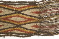 Sash Closeup. Woven sash showing a diamond pattern in red, black and yellow, with white beads on the edge. The proper left shows the ending fringe of the sash.